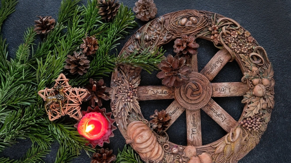 Wheel of the Year with carved seasonal names and natural decorations, surrounded by pinecones, greenery, a red candle, and a wicker star ornament on a dark background