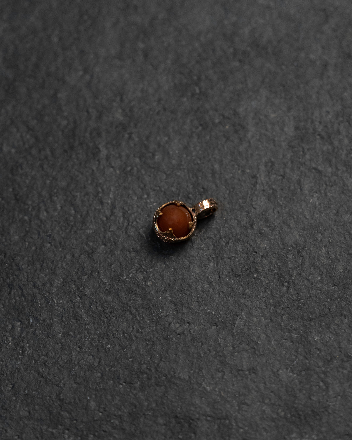 A small round pendant or charm with a warm brown carnelian stone in a textured gold-toned bezel setting, photographed on a dark textured surface.