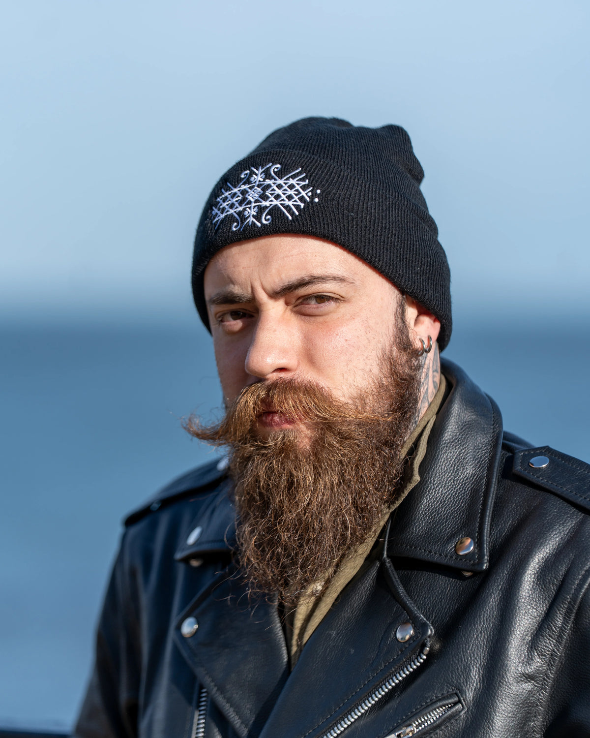 A close-up portrait of a man with a thick brown beard wearing a black knit beanie with a white embroidered bindrune design and a black leather motorcycle jacket, set against a soft blue sky and water background.
