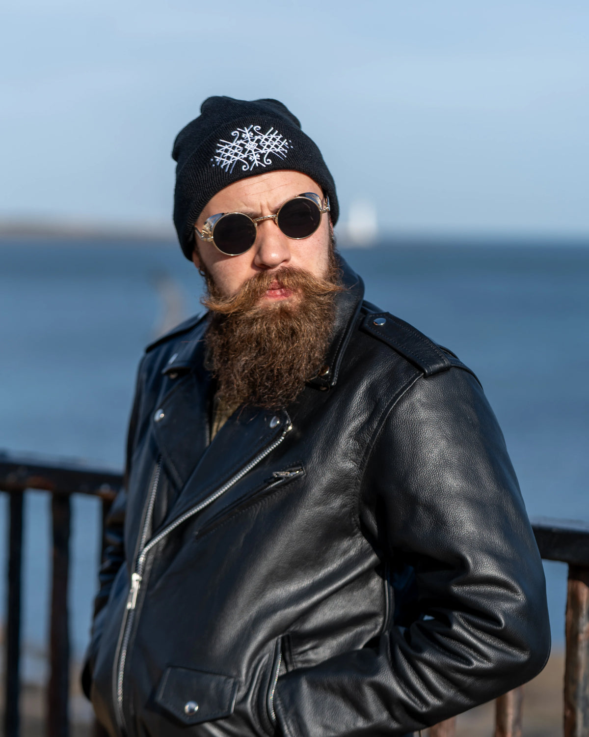 A portrait of a man with a thick brown beard wearing a black knit beanie with a white embroidered bindrune design and a black leather motorcycle jacket, set against a soft blue sky and water background.