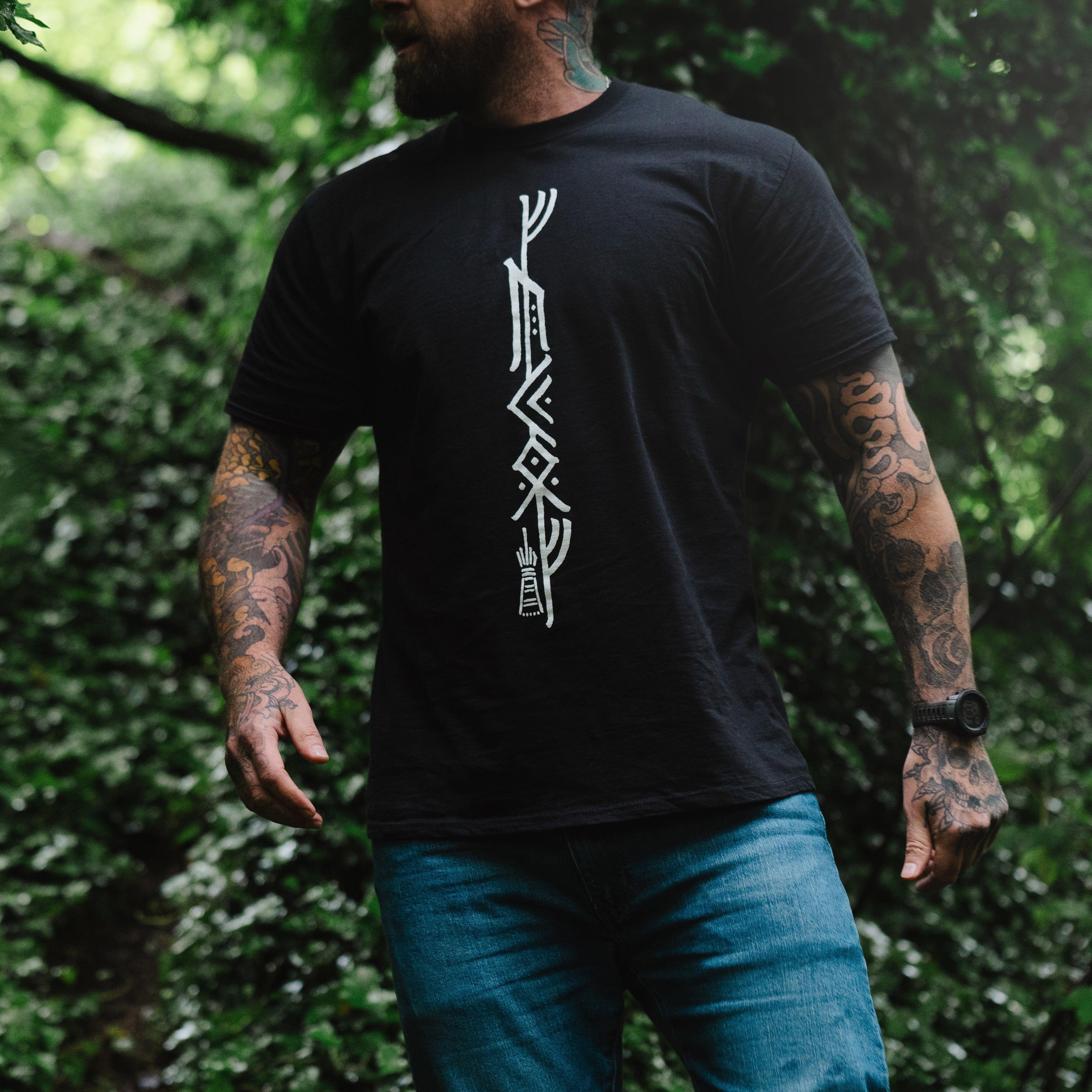 Man in forest wearing black t-shirt with Norse runes, showcasing Viking-inspired apparel and tattoo art.