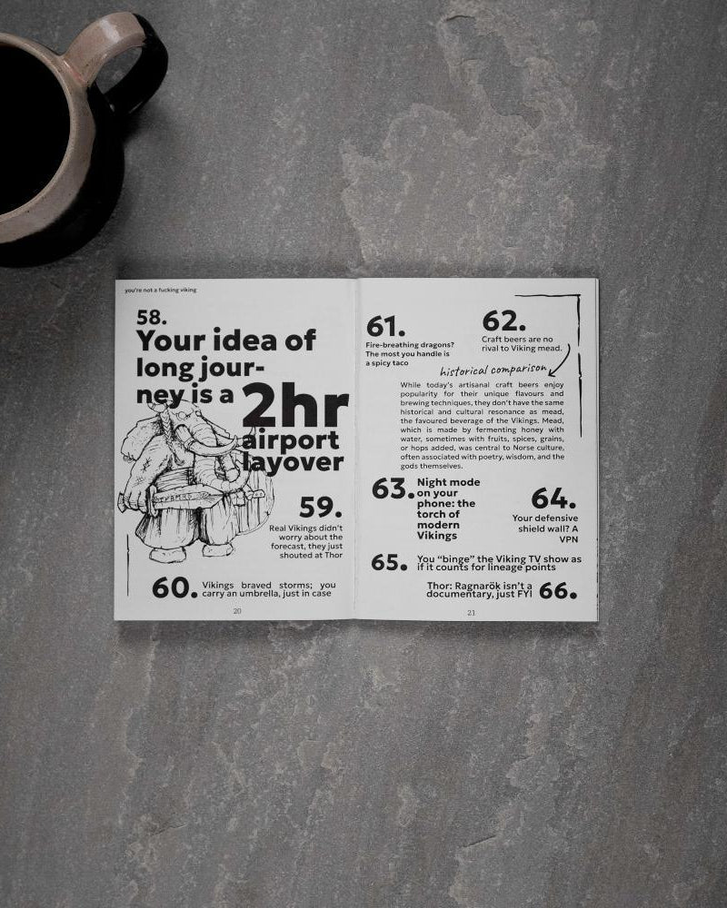 Open Viking art book with humorous Norse facts and illustrations, paired with coffee mug on stone surface