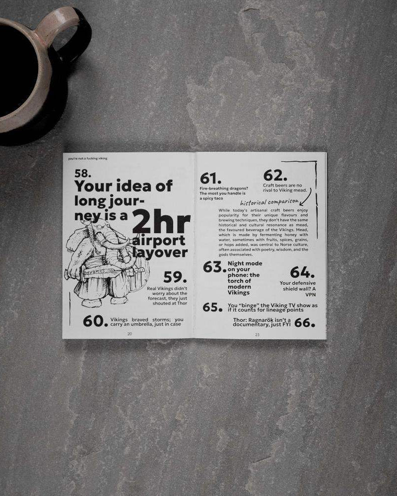 Open Viking art book with humorous Norse facts and illustrations, paired with coffee mug on stone surface