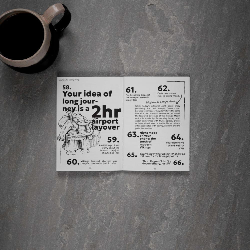Open Viking art book with humorous Norse facts and illustrations, paired with coffee mug on stone surface