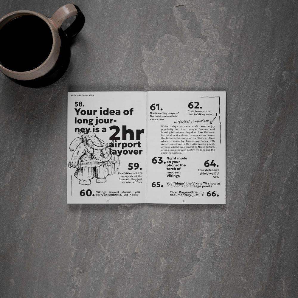 Open Viking art book with humorous Norse facts and illustrations, paired with coffee mug on stone surface