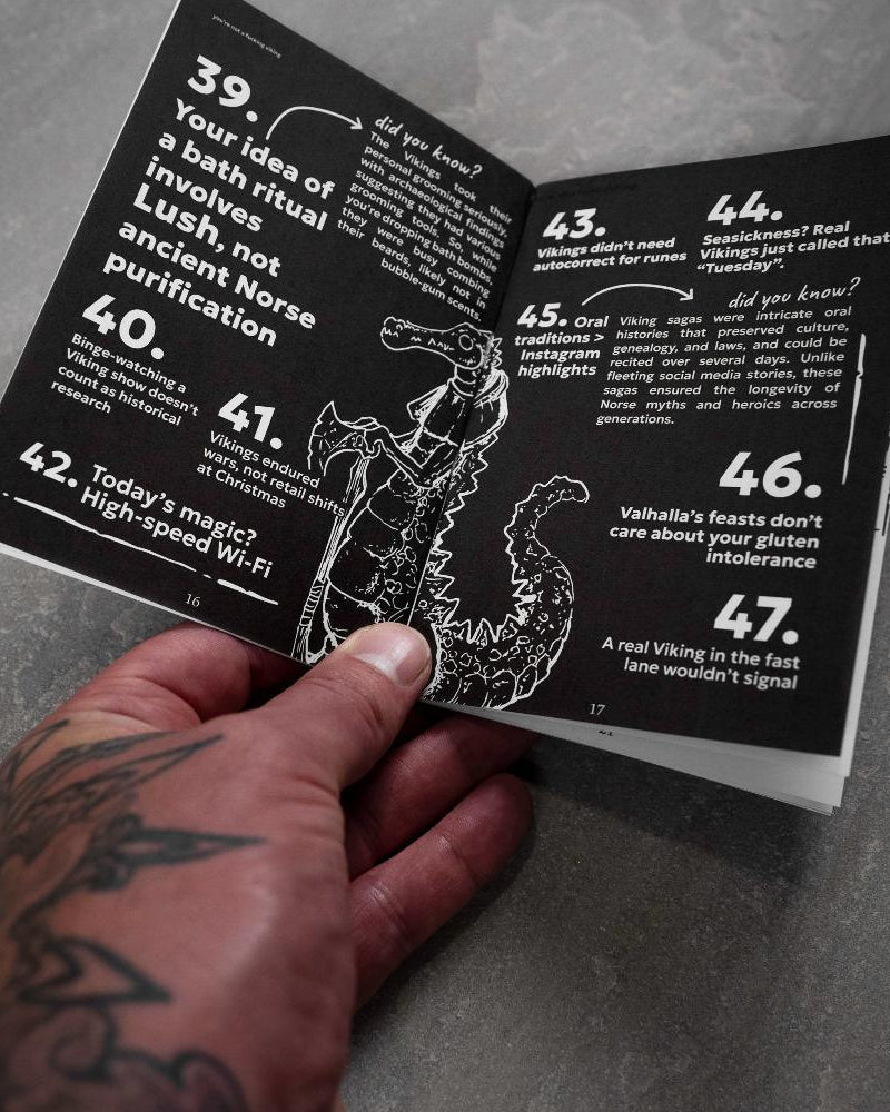 Hand holding Norse myth book with Viking facts and dragon illustration, tattooed arm visible