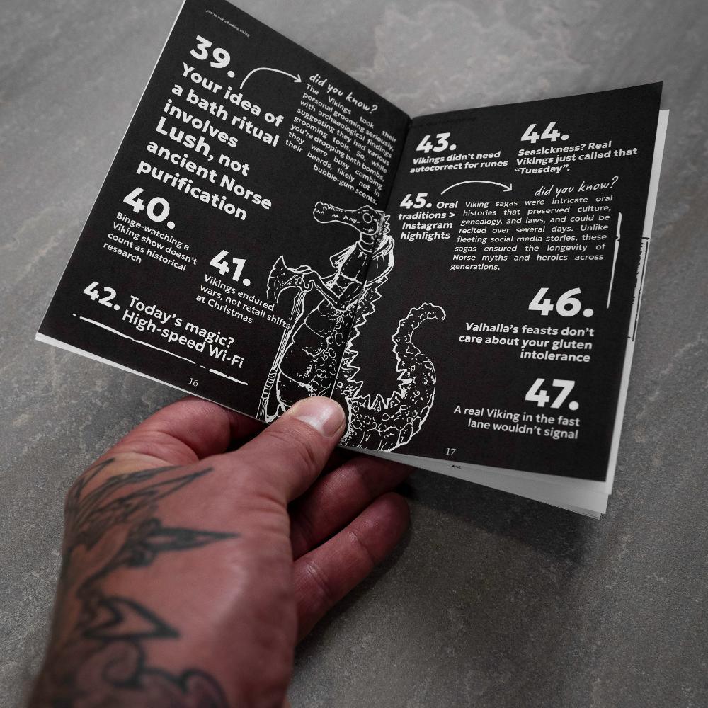 Hand holding Norse myth book with Viking facts and dragon illustration, tattooed arm visible