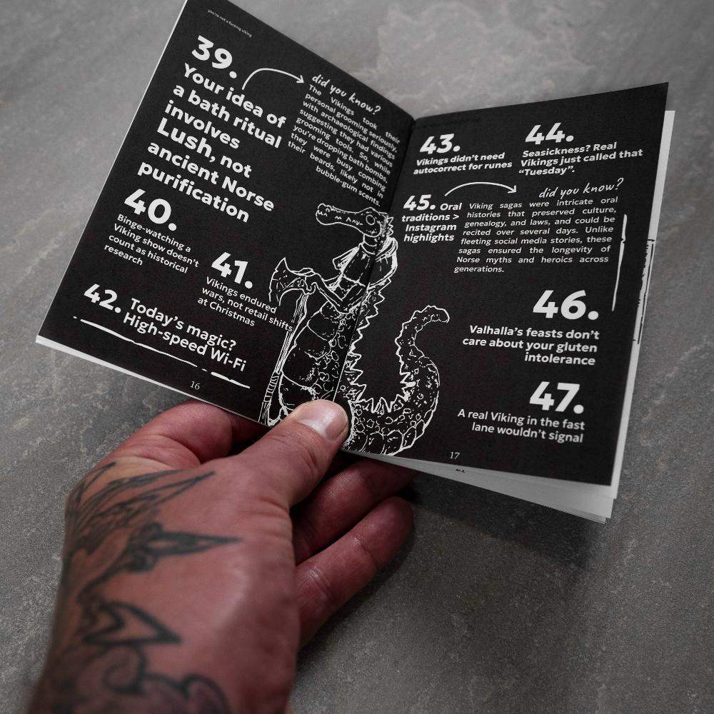 Hand holding Norse myth book with Viking facts and dragon illustration, tattooed arm visible