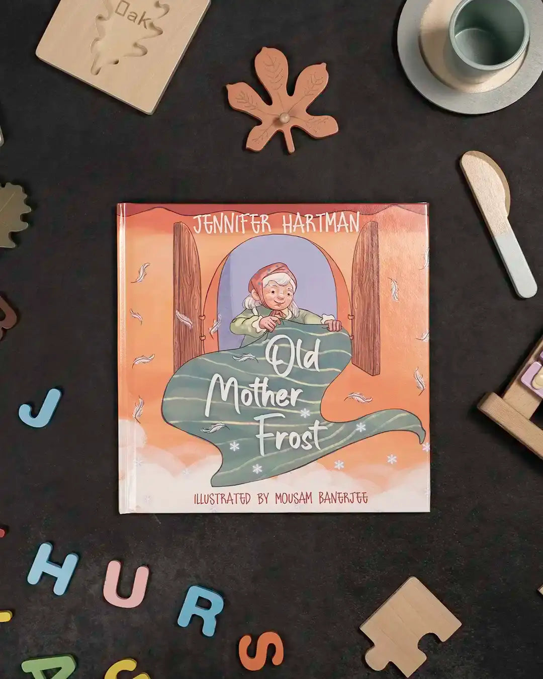 Old Mother Frost children's book by Jennifer Hartman, illustrated with cozy fairy tale art, surrounded by wooden toys and letters.