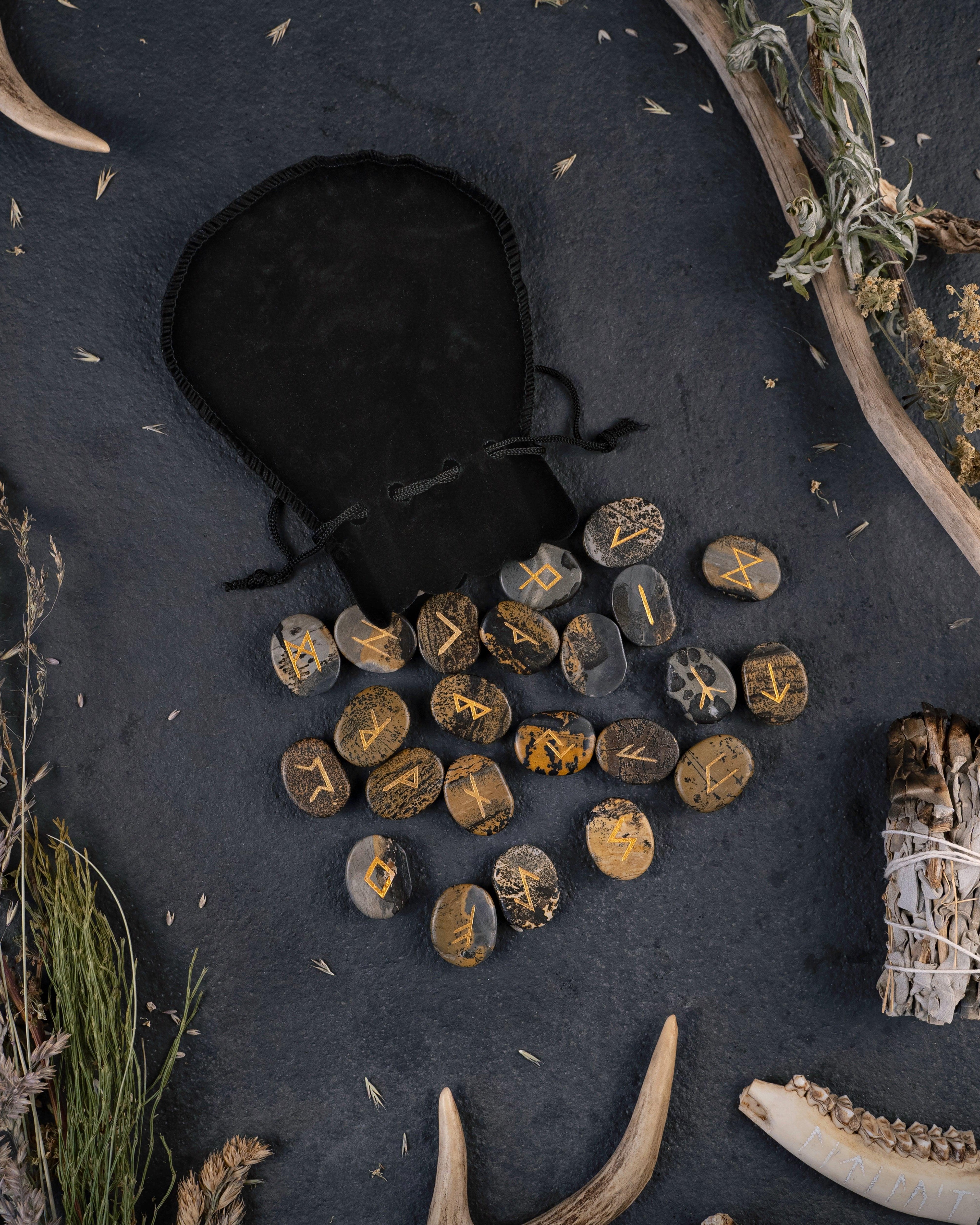Norse rune stones with golden symbols, antlers, dried herbs, and a black pouch on dark surface