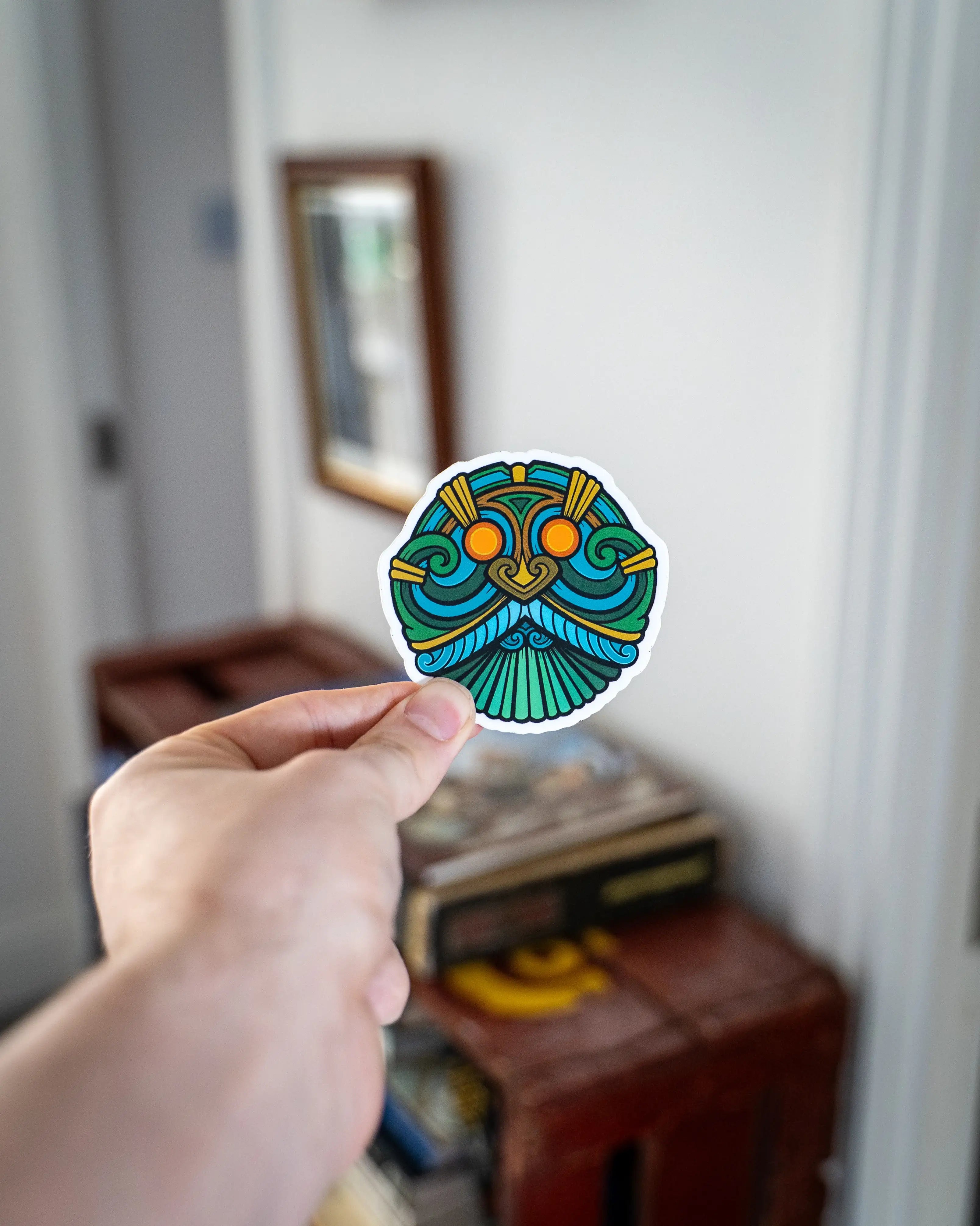 Hand holding vibrant Norse-inspired sticker with Viking mask design, featuring teal, green, and gold tribal patterns.