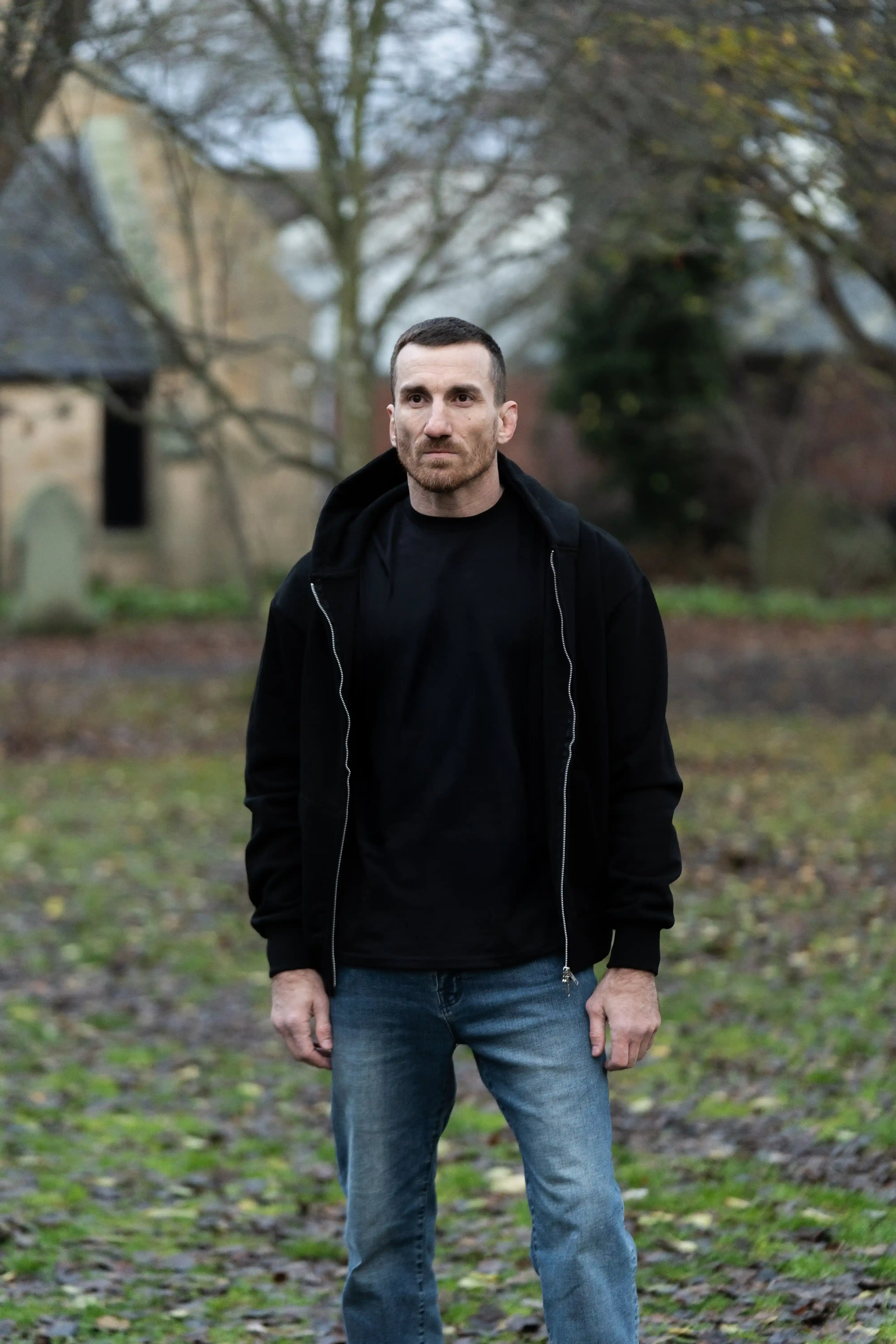 Man in black hoodie and jeans standing outdoors near a rustic building