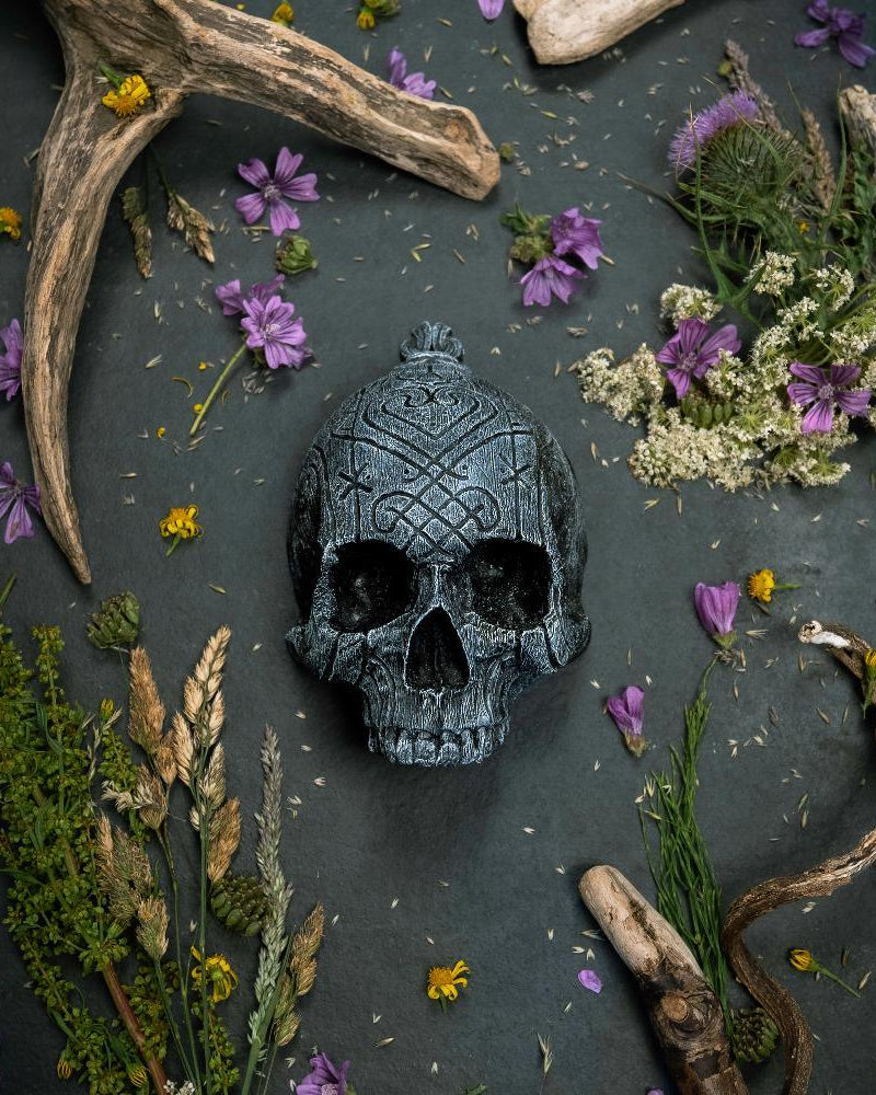 Carved Norse skull with Viking runes, surrounded by antlers, dried flowers, and herbs on dark stone.
