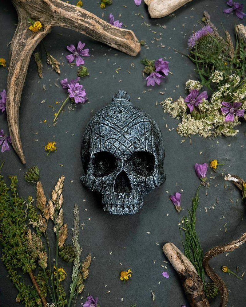 Carved Norse skull with Viking runes, surrounded by antlers, dried flowers, and herbs on dark stone.