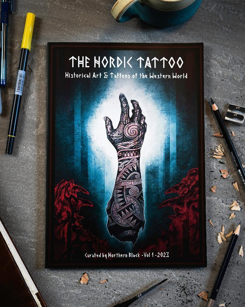 The Nordic Tattoo art book with Viking-inspired hand tattoo design, surrounded by pens and pencils on concrete.