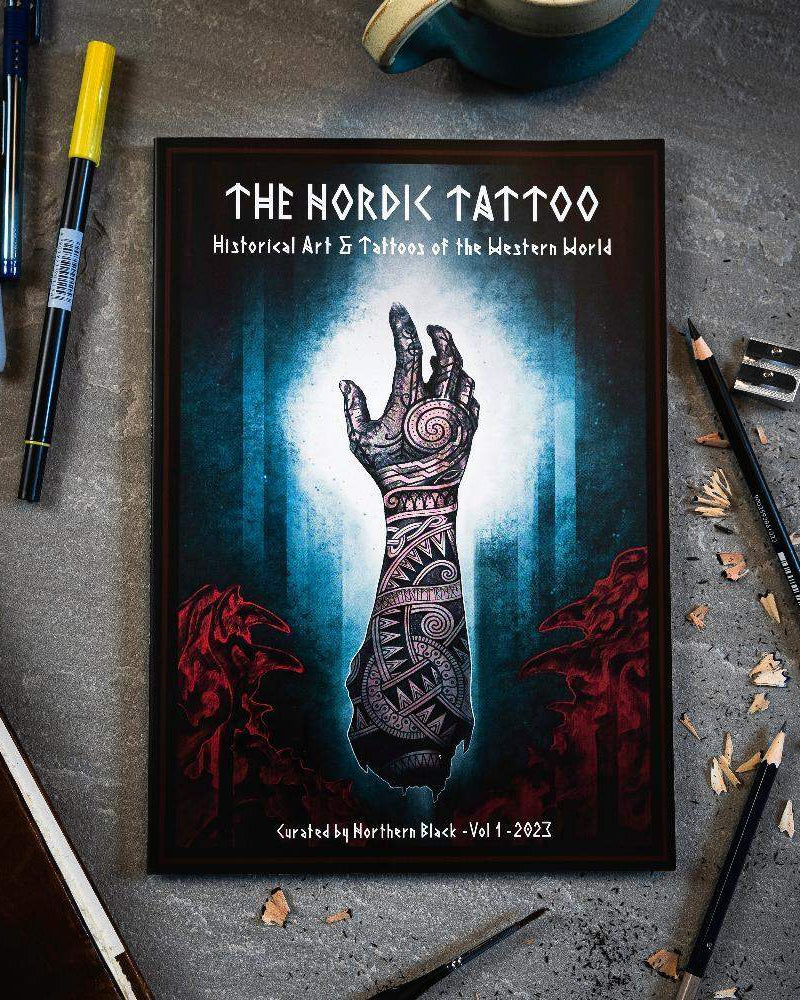 The Nordic Tattoo art book with Viking-inspired hand tattoo design, surrounded by pens and pencils on concrete.