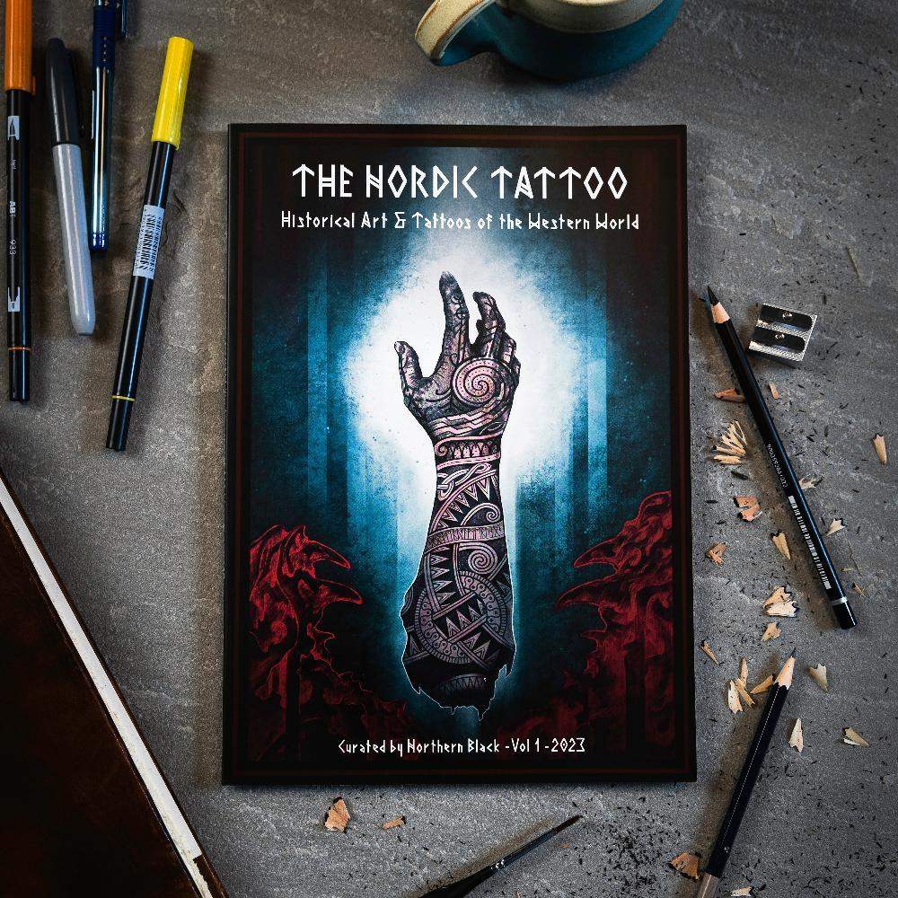 The Nordic Tattoo art book with Viking-inspired hand tattoo design, surrounded by pens and pencils on concrete.