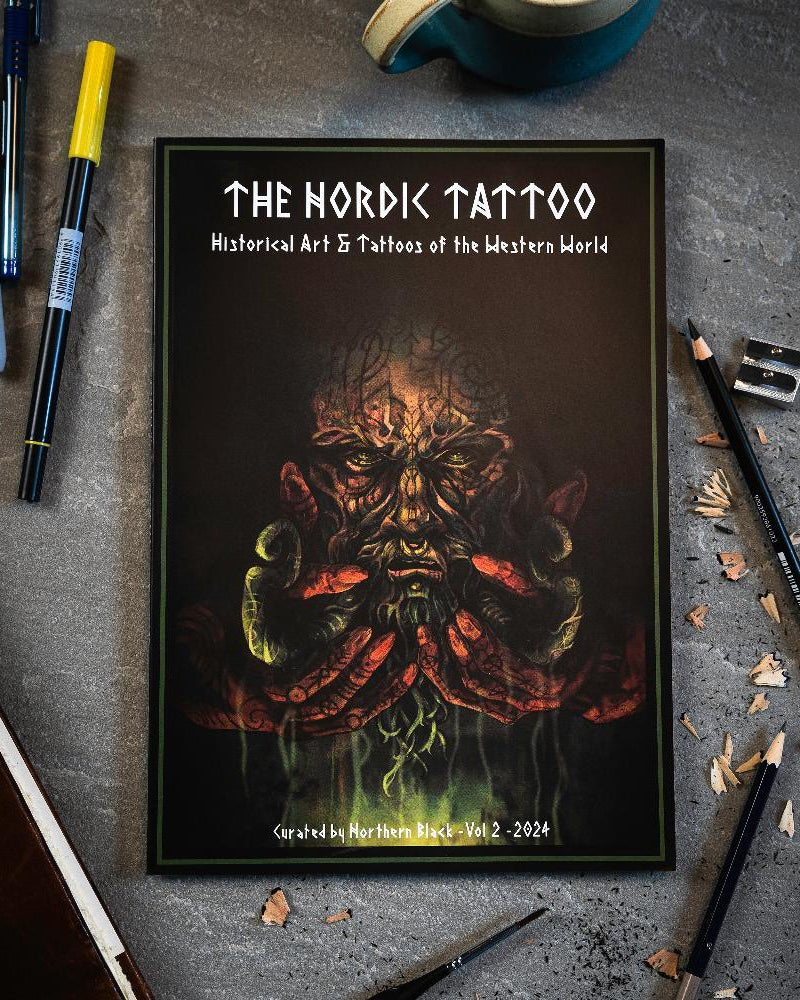 The Nordic Tattoo art book with Viking-inspired cover, surrounded by pens and pencils on a desk