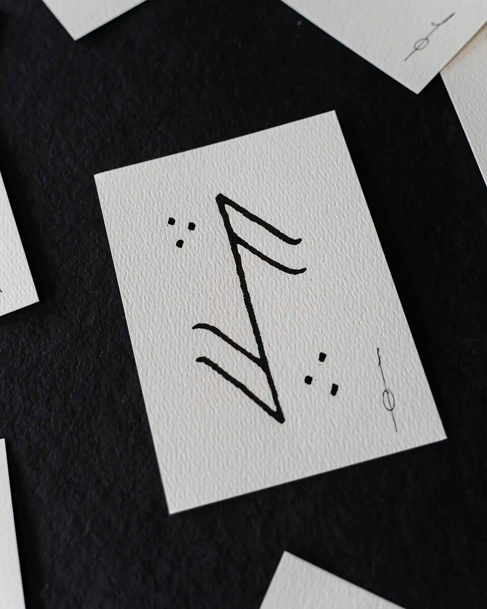 A close-up of a white textured card featuring a black hand-drawn rune symbol, surrounded by scattered matching white cards on a dark background.
