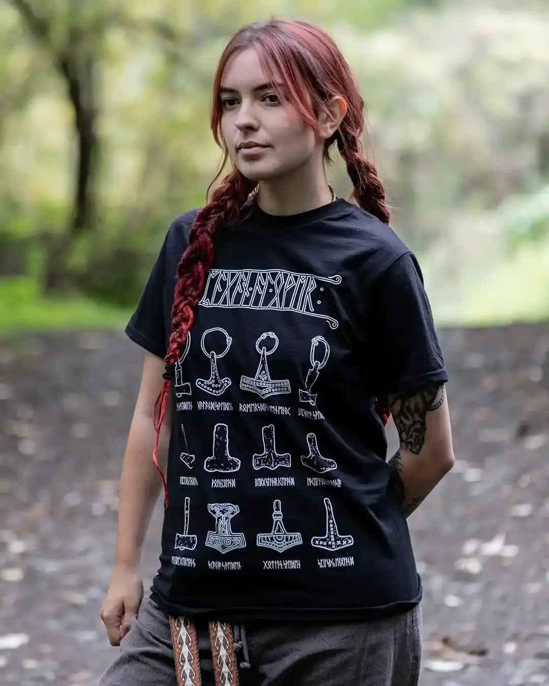 Woman wearing Norse-inspired black t-shirt with Viking hammer designs and runes, outdoors in forest setting