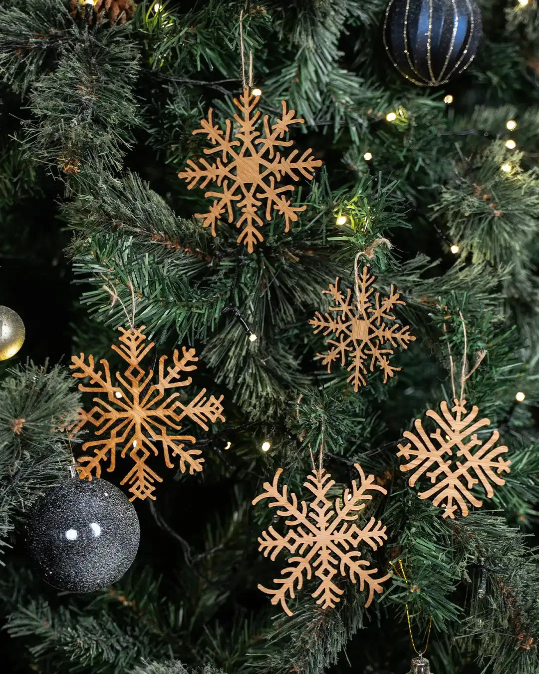 Wooden snowflake ornaments hanging on a Christmas tree with twinkling lights and black baubles.
