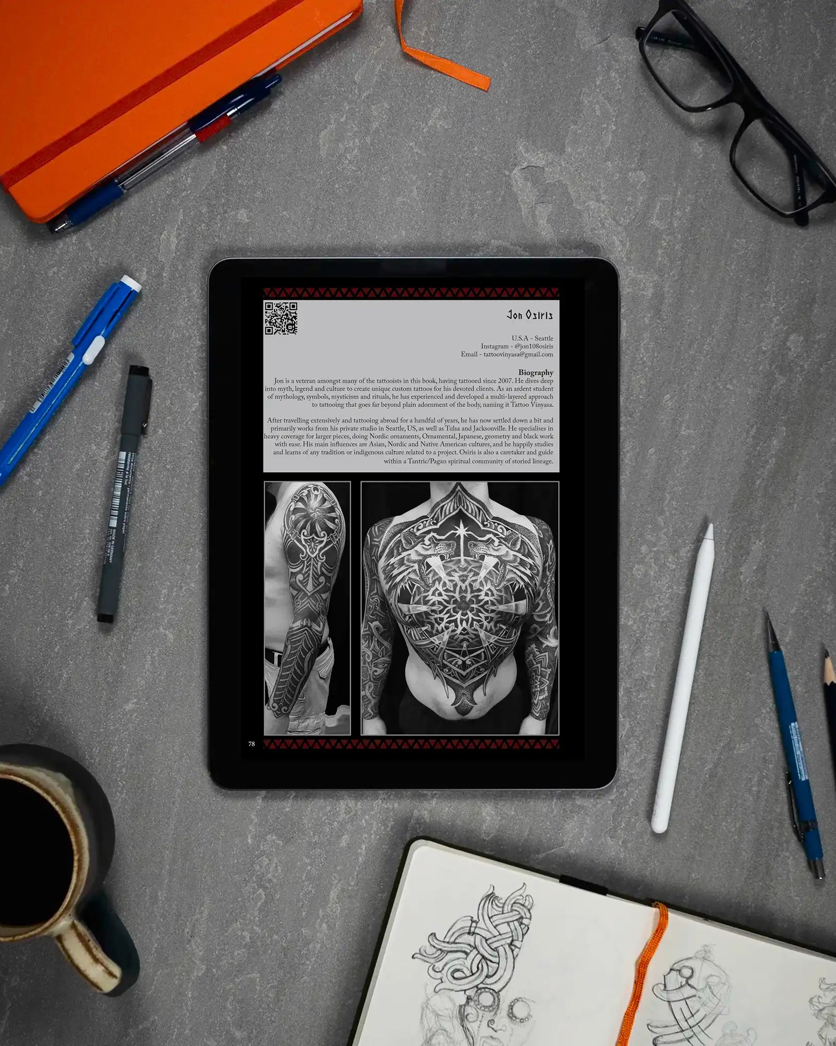 Viking tattoo art book on tablet with Norse designs, sketchbook, pens, and coffee on desk