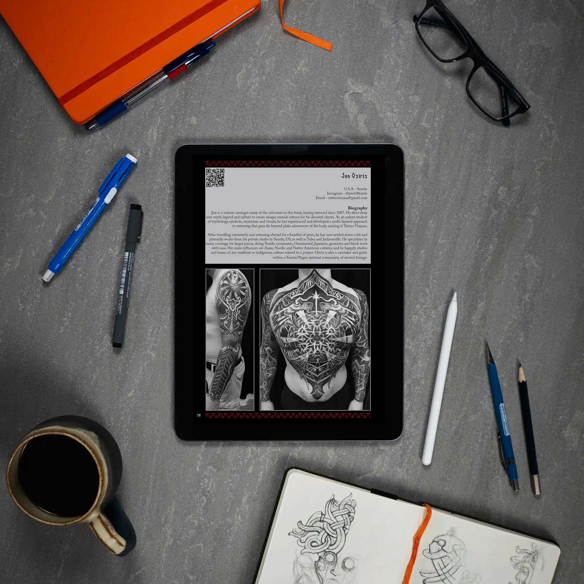 Viking tattoo art book on tablet with Norse designs, sketchbook, pens, and coffee on desk