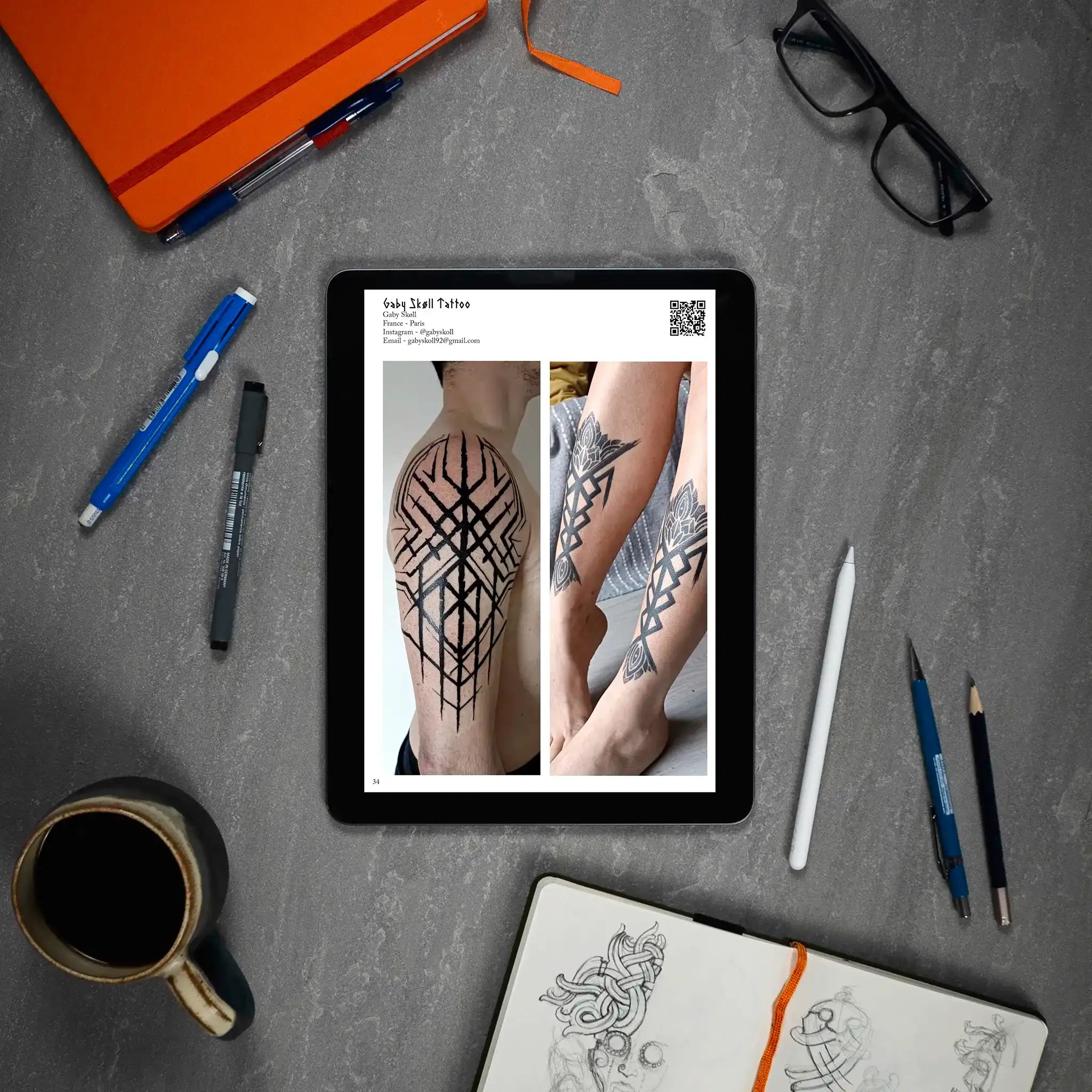 Viking tattoo designs on tablet: geometric Norse patterns on arm and legs, surrounded by sketchbook, pens, coffee