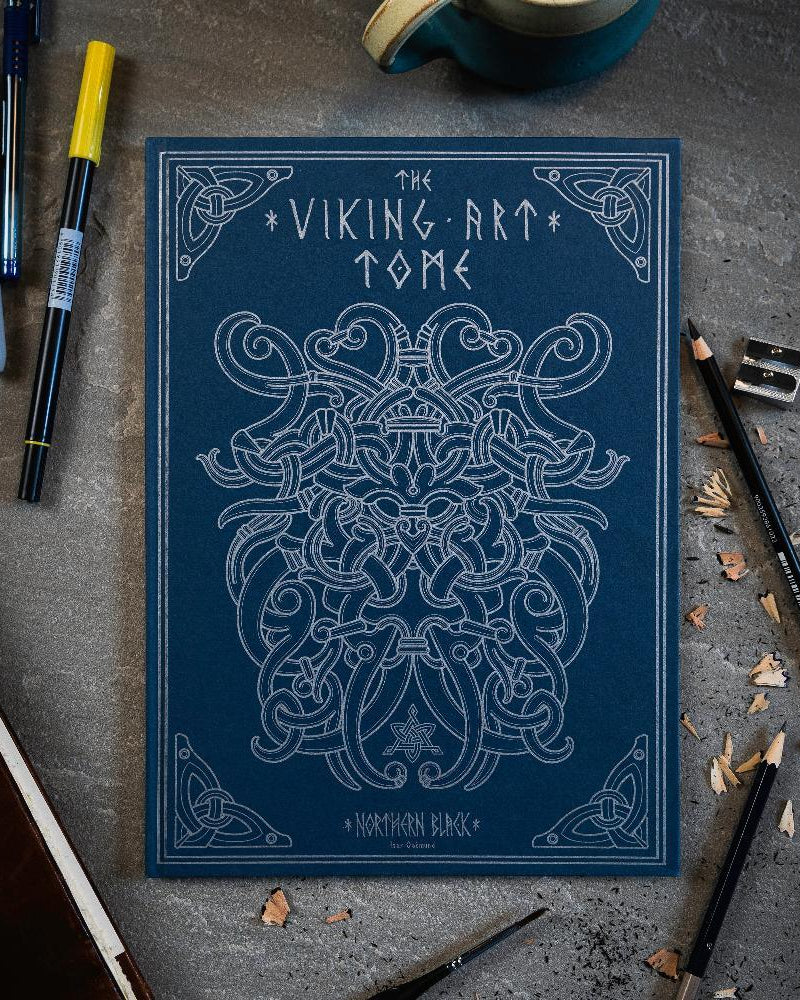 The Viking Art Tome with intricate Norse designs, surrounded by pens and pencils on a textured surface.