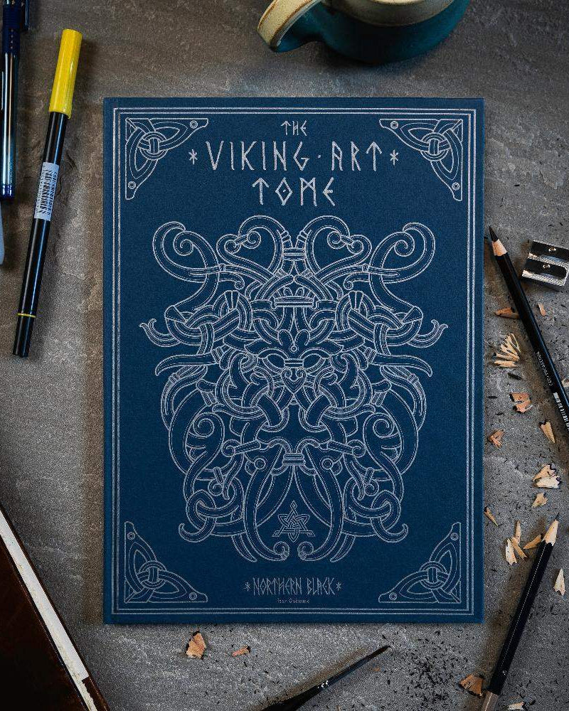 The Viking Art Tome with intricate Norse designs, surrounded by pens and pencils on a textured surface.