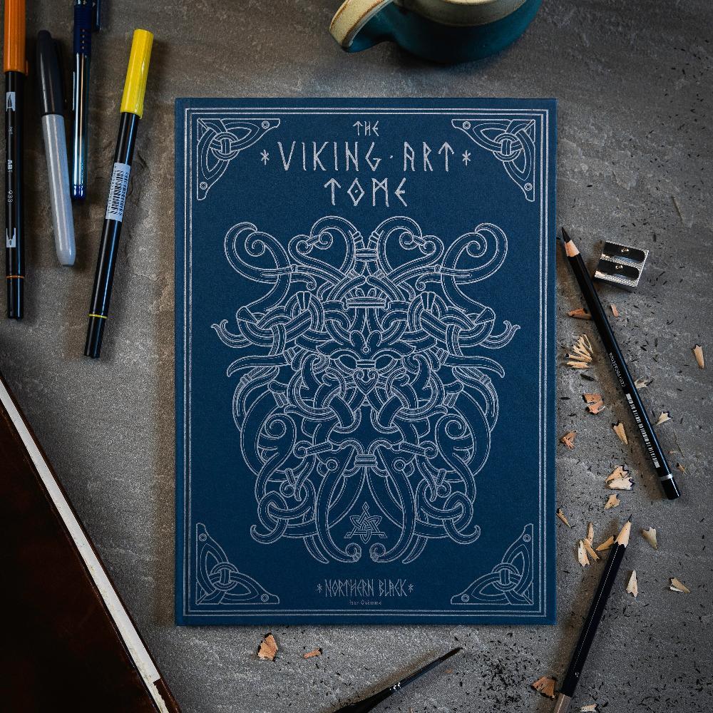 The Viking Art Tome with intricate Norse designs, surrounded by pens and pencils on a textured surface.