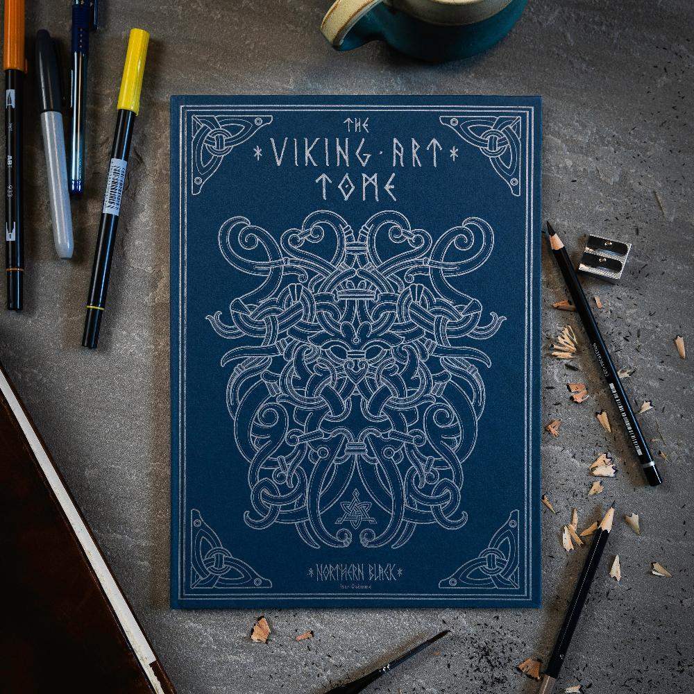 The Viking Art Tome with intricate Norse designs, surrounded by pens and pencils on a textured surface.