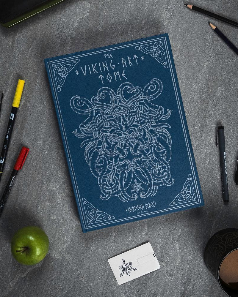 The Viking Art Tome with Norse knotwork design, surrounded by pens, pencils, apple, and mug on gray surface