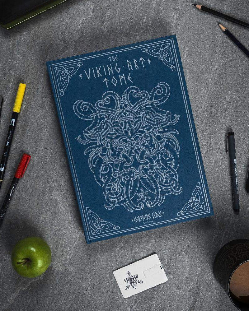 The Viking Art Tome with Norse knotwork design, surrounded by pens, pencils, apple, and mug on gray surface