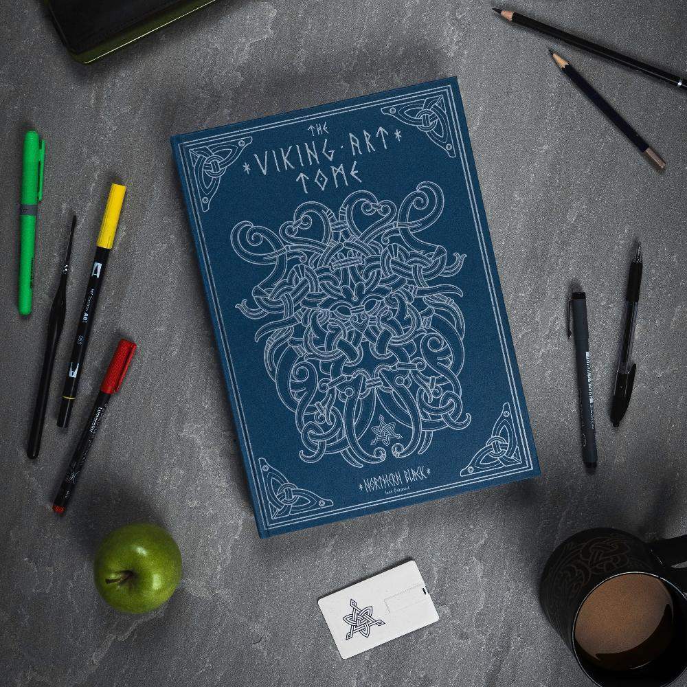 The Viking Art Tome with Norse knotwork design, surrounded by pens, pencils, apple, and mug on gray surface