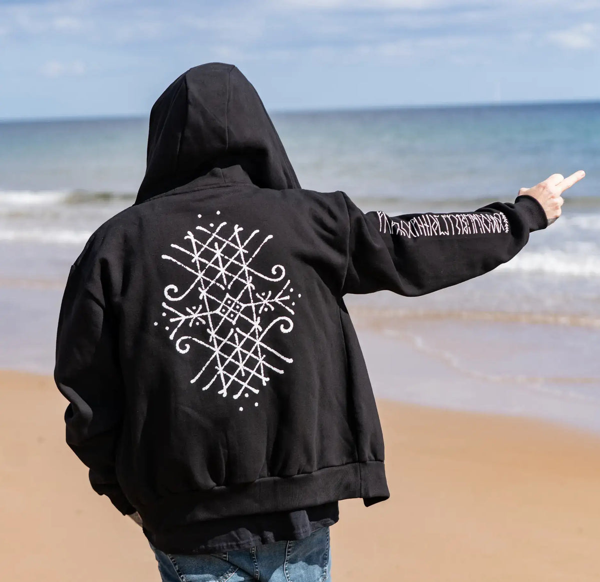 Black hoodie with Viking rune design on back, worn on beach, Norse-inspired apparel