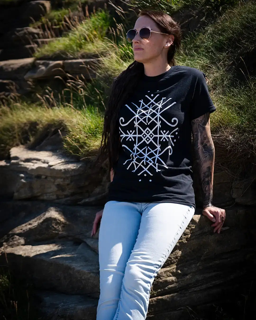 Woman wearing Norse-inspired black t-shirt with white Viking rune design, sitting on rocky cliffside