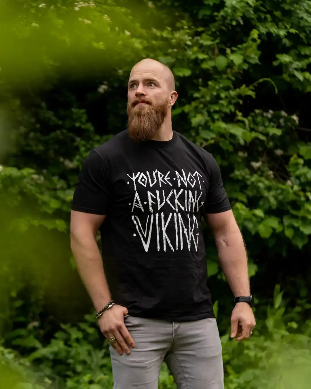 Man wearing bold Viking-themed black t-shirt with edgy white text, standing outdoors in green foliage.