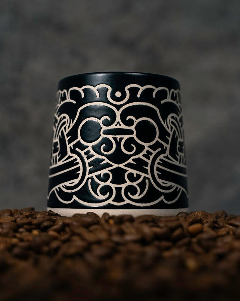 Black ceramic mug with intricate Norse-inspired Viking art design, set on coffee beans