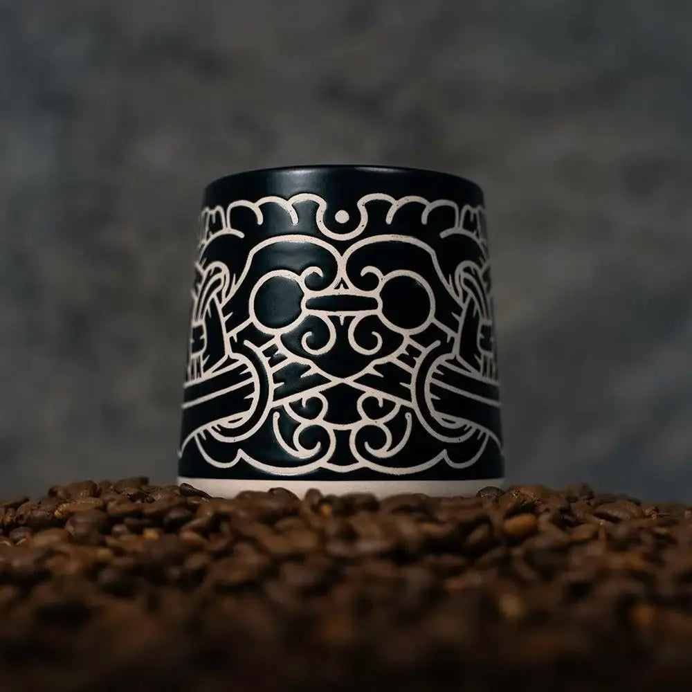 Black ceramic mug with intricate Norse-inspired Viking art design, set on coffee beans