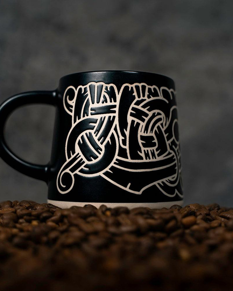 Black mug with Norse knotwork design, surrounded by coffee beans — Viking-inspired art