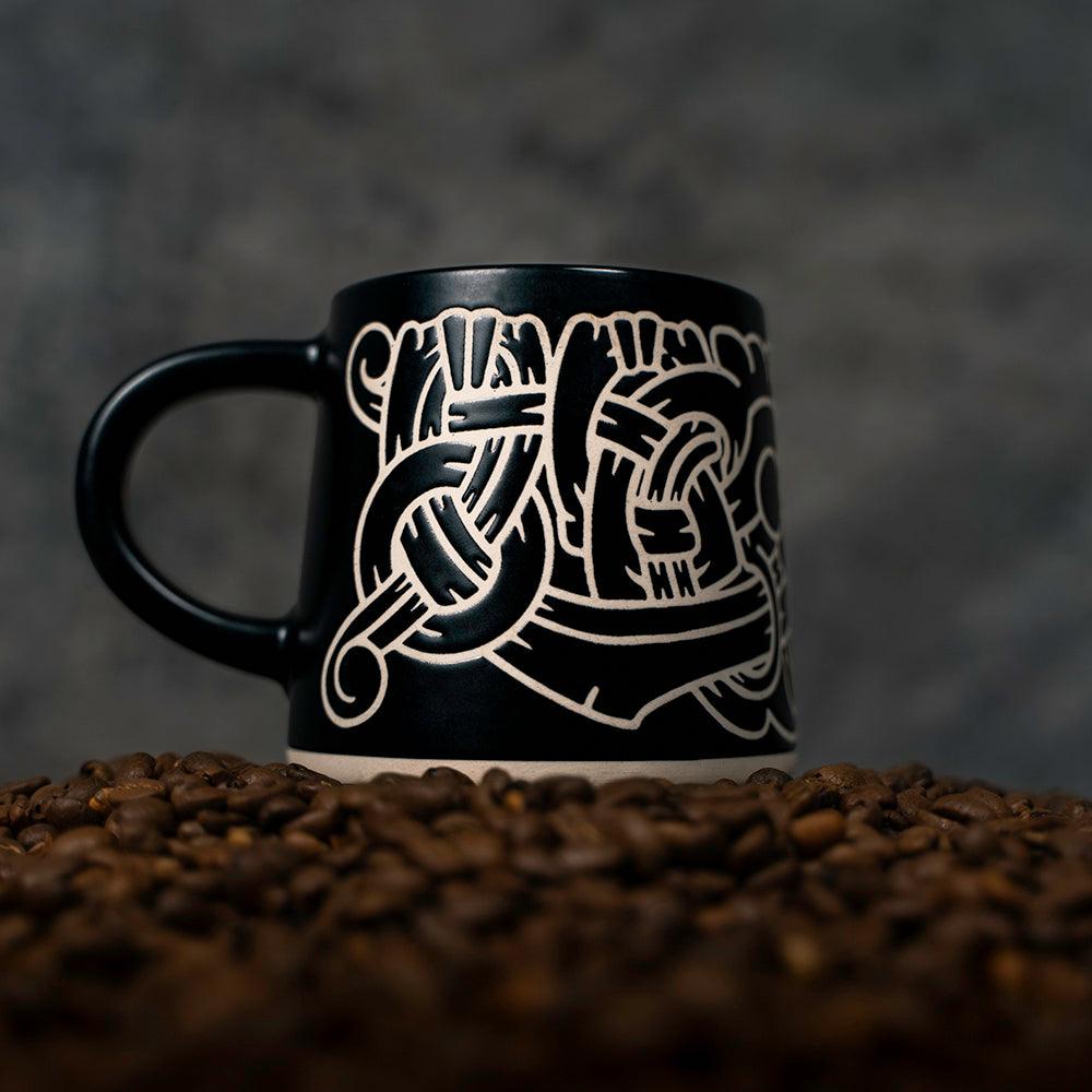 Black mug with Norse knotwork design, surrounded by coffee beans — Viking-inspired art