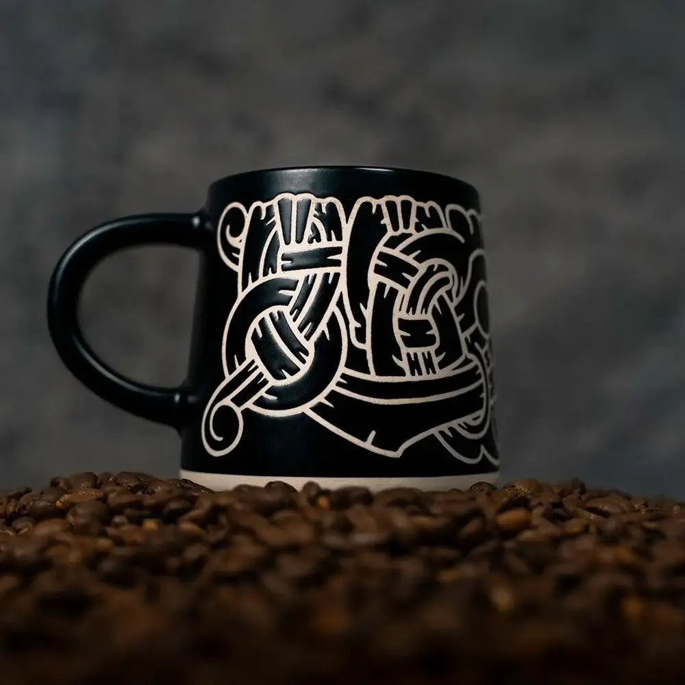Black mug with Norse knotwork design, surrounded by coffee beans — Viking-inspired art