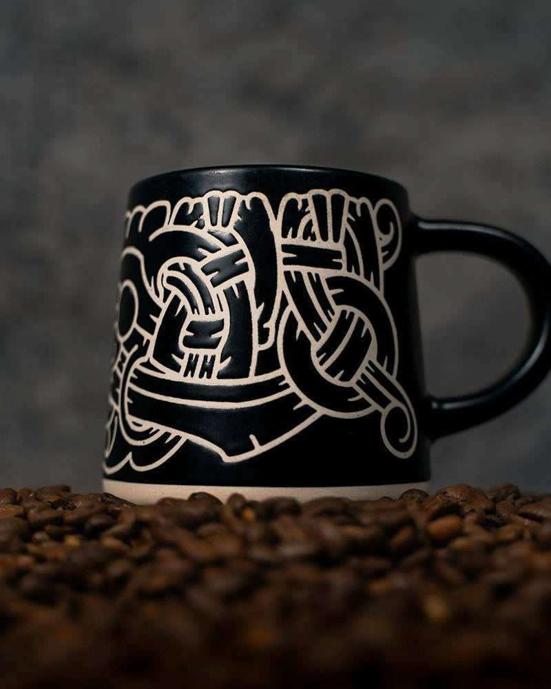 Black mug with white Norse knotwork design, set on coffee beans — Viking-inspired ceramic art.