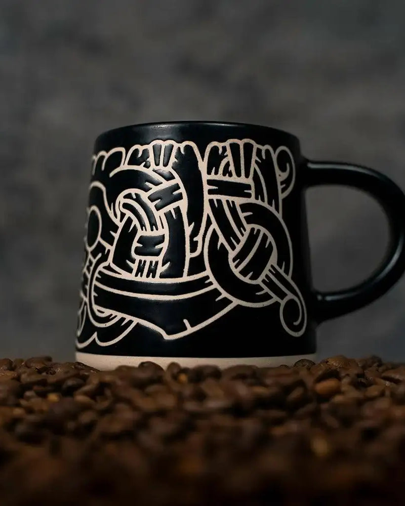 Black mug with white Norse knotwork design, set on coffee beans — Viking-inspired ceramic art.