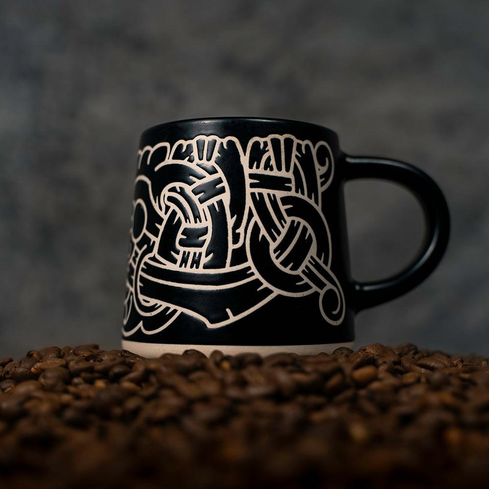 Black mug with white Norse knotwork design, set on coffee beans — Viking-inspired ceramic art.
