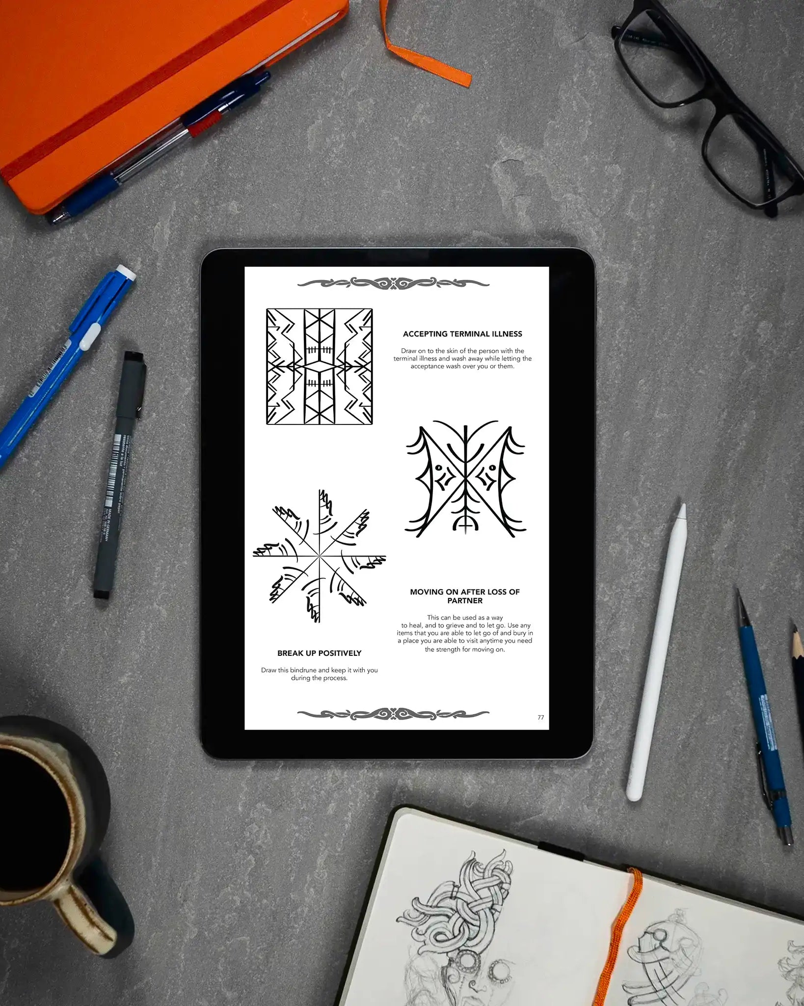 Tablet displaying Norse-inspired bindrune designs for healing and acceptance, surrounded by sketchbook, pens, and coffee.