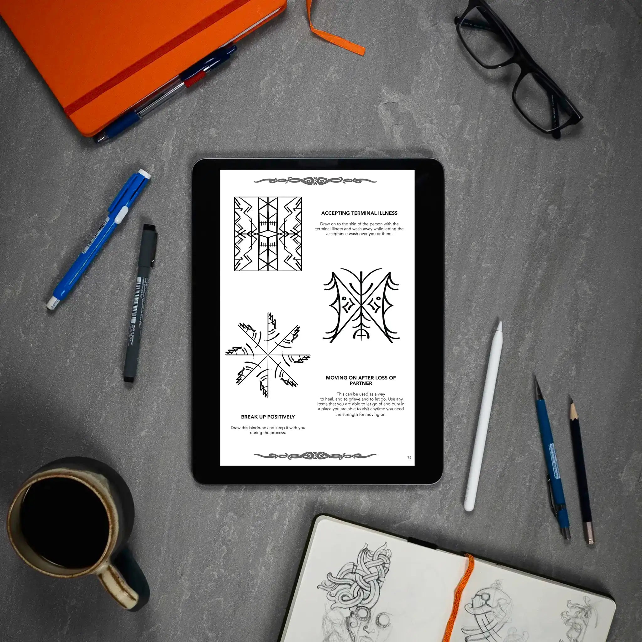 Tablet displaying Norse-inspired bindrune designs for healing and acceptance, surrounded by sketchbook, pens, and coffee.