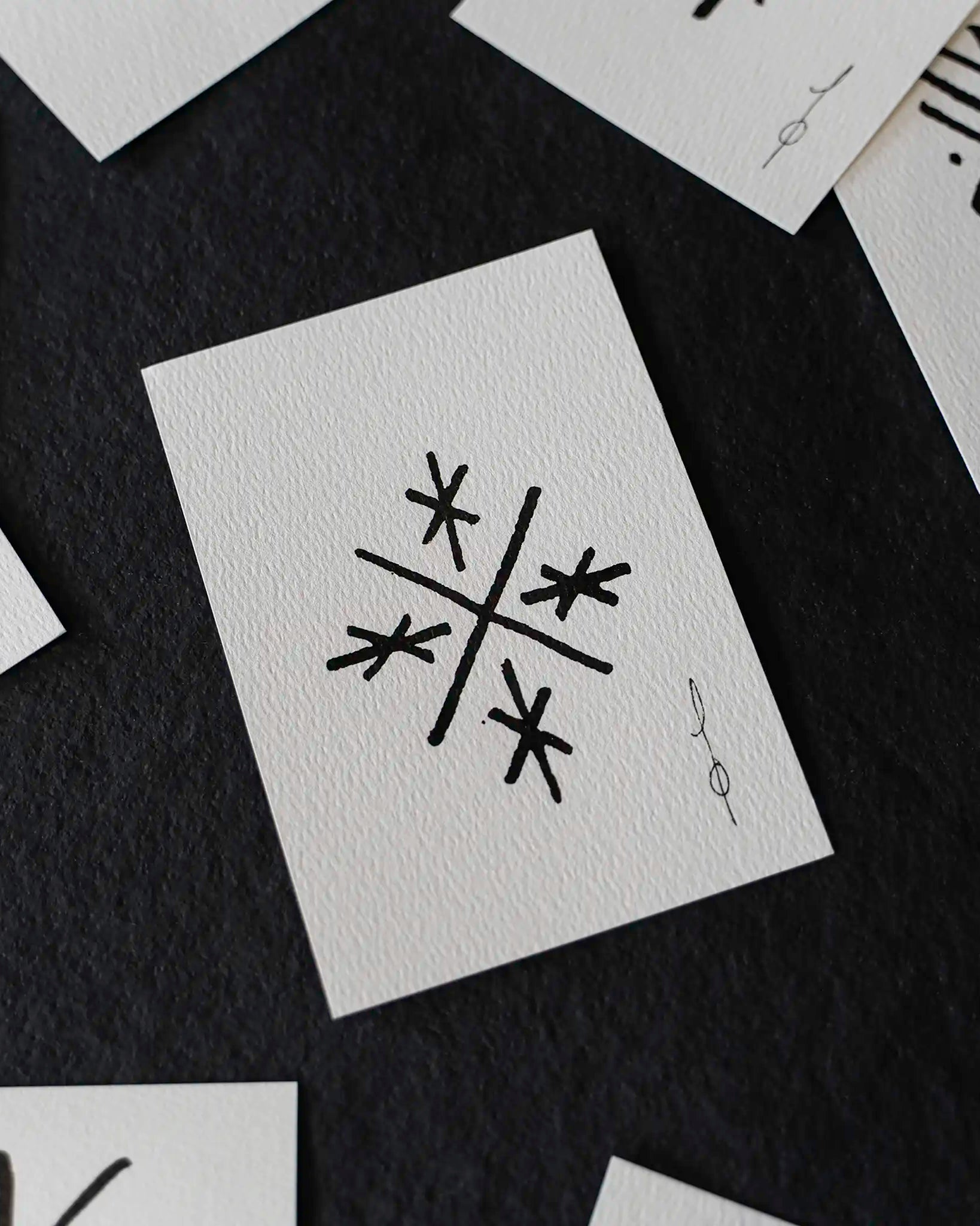 A close-up flat-lay of several white textured cards scattered on a dark charcoal background. The central card shows a black abstract geometric symbol resembling a stylized snowflake or starburst, with a small vertical signature-like mark beside it.