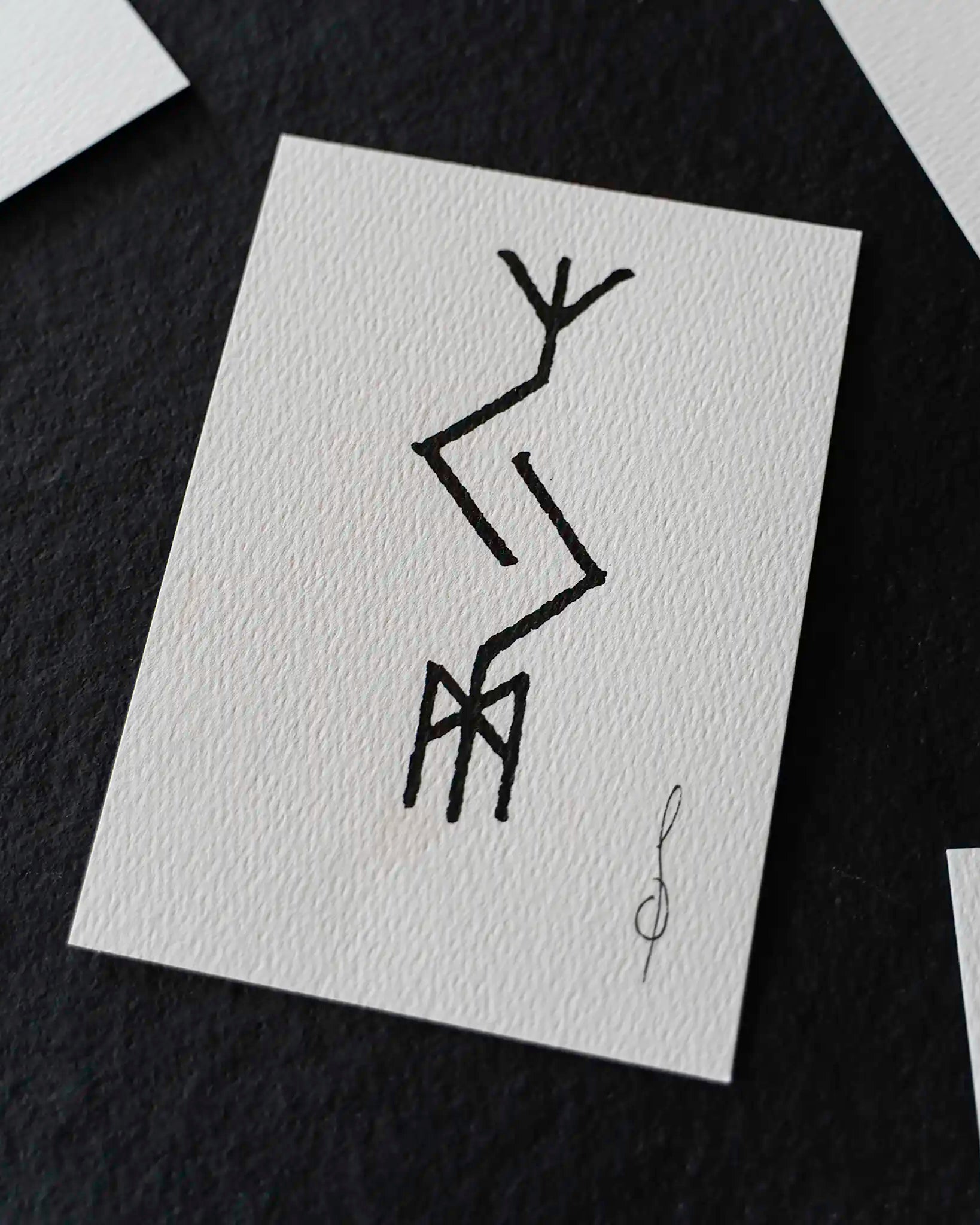 A square white art print with a black abstract line drawing, displayed on a dark textured surface with other partial prints around it.
